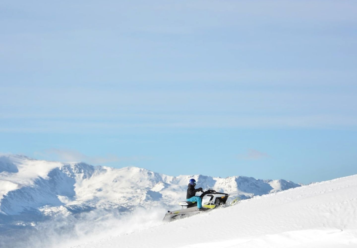 person snowmobiling up a mountain with a mountain range in the background