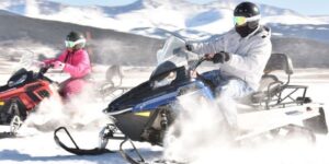 two people riding snowmobiles with mountains and trees in the background